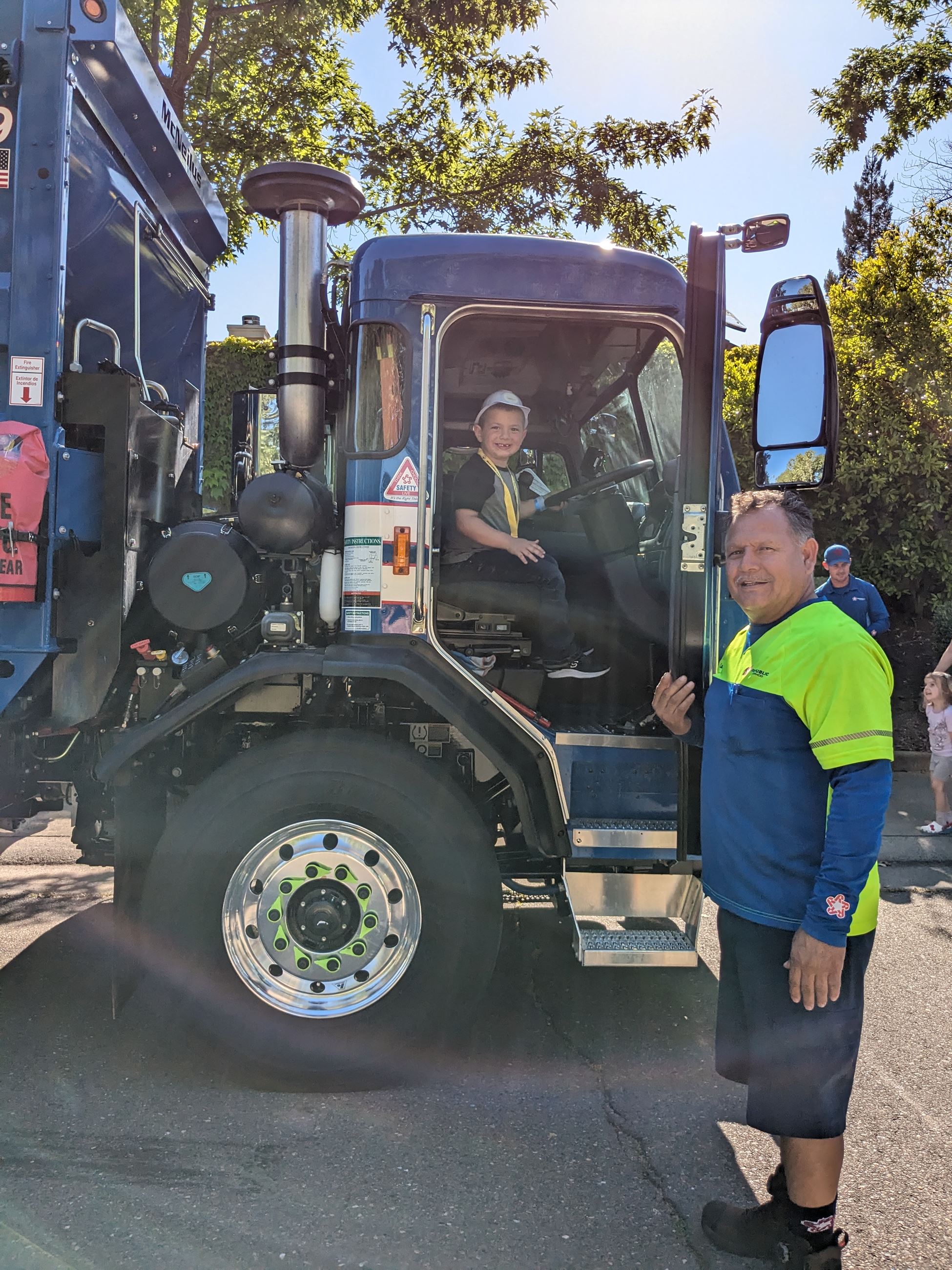 A child poses in the front seat of a garbage truck while an adult stands at the door.