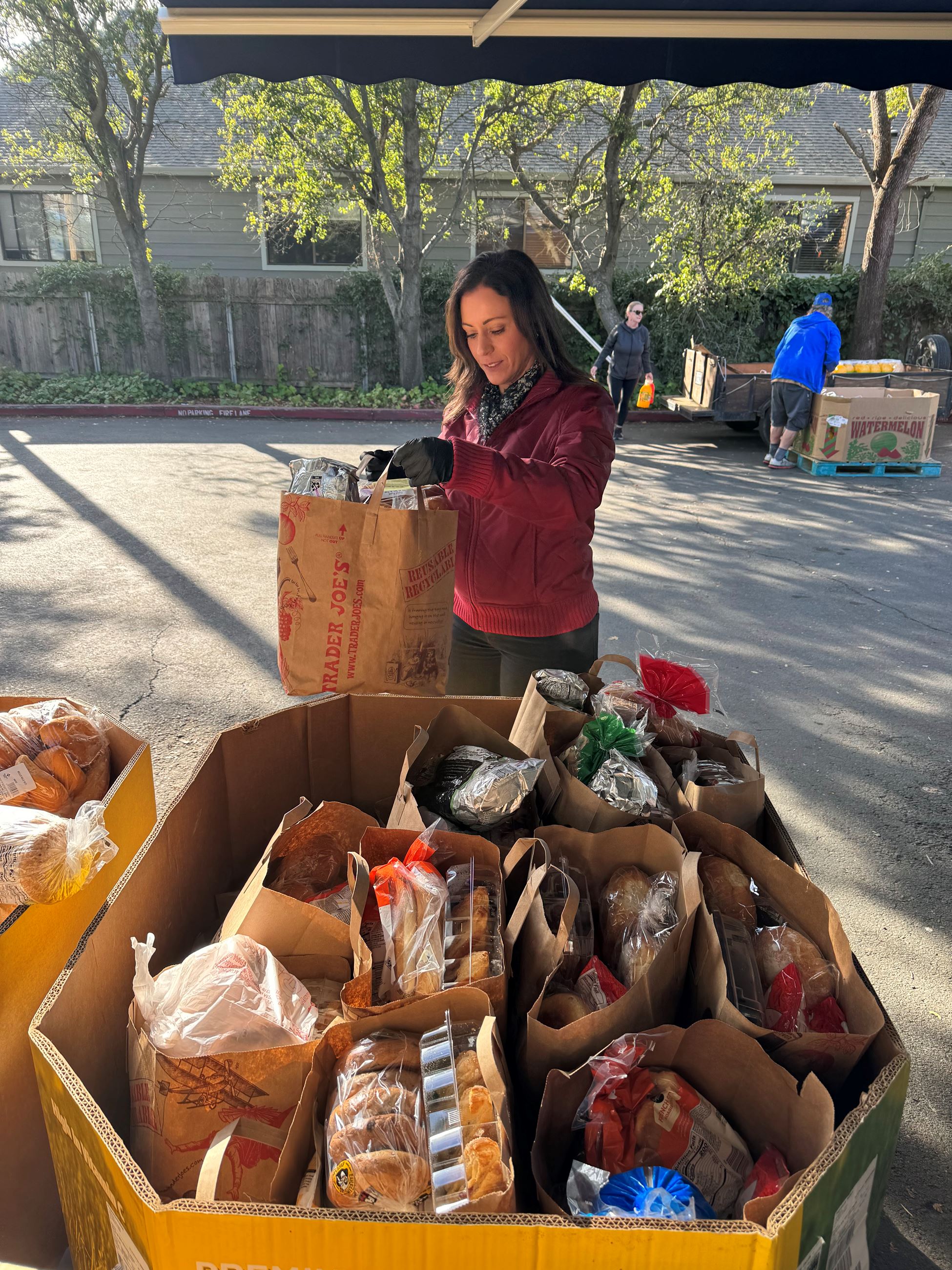 A woman holds a brown bag full of food, food items sit in a large cardboard bin in front of her.